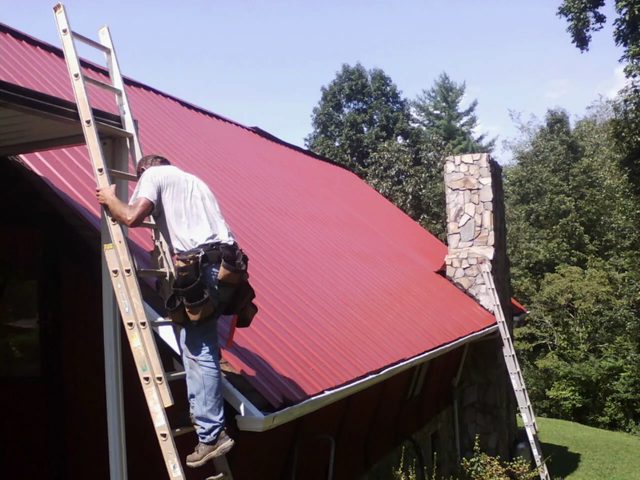 Roofer on a ladder beside a red metal roof and stone chimney — Soffit & Fascia Repair by Roofing Brothers in Jbsa Ft Sam Houston